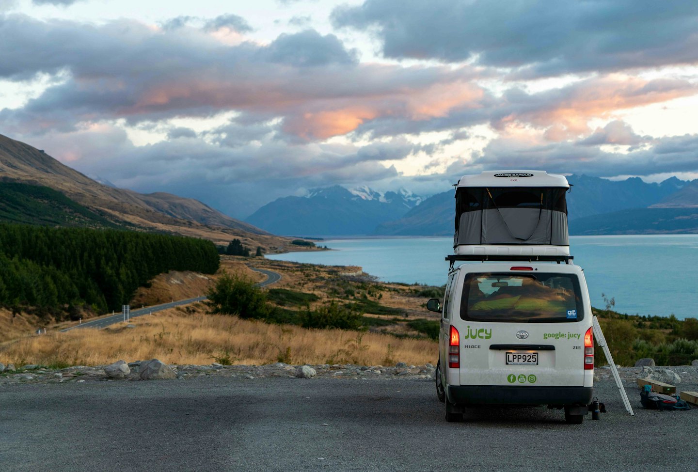 Furgoneta camper blanca con tienda de techo aparcada junto a un lago de montaña.